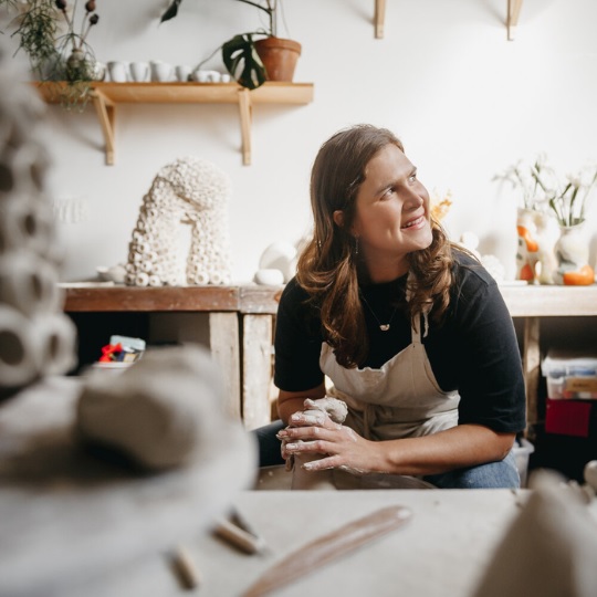 Photo of Milly Dent in her studio, she sits at a clay wheel bent over a piece in progress, her hands have whiute clay on them as she forms a piece with her hands, she has long sandy blond hair, a black ltshirt that covers her uper arms and jeans, she looks off to the side smiling happily with a cheeky expression, the the foreground are out of focus clay pieces and in the background are shelves with ceramic pieces, plants and tools